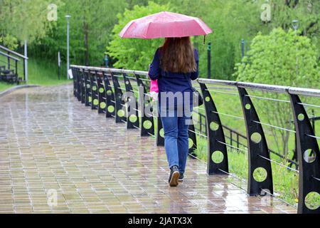 Starker Regen, Frau mit Regenschirm läuft im grünen Park am nassen Bürgersteig Stockfoto