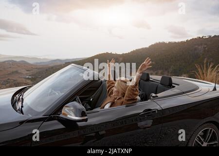Junge, fröhlich lächelnde Frau, die im Cabriolet am Berghang unterwegs ist, Seitenansicht Stockfoto