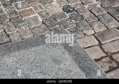 Ecke eines Fußgängerwegs aus Granit-Bordstein und rechteckigen Steinfliesen aus der Nähe, im Hintergrund eine Straße mit grob geschnittenen Pflastersteinen gepflastert Stockfoto