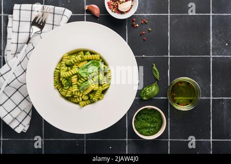 Italienische Fusilli Pasta mit Pesto. Fusilli-Pasta mit Basilikumpesto und Kräutern, Parmesankäse, Basilikum und Knoblauch auf weißem Teller auf Arbeitsplatte aus schwarzem Stein Stockfoto