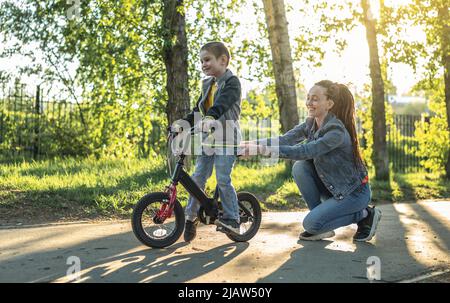 Mama hilft einem kleinen Jungen, ein Fahrrad mit zwei Rädern im Park zu fahren. Ein angenehmer Sommersporturlaub für Kinder. Stockfoto