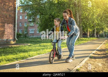 Mama hilft einem kleinen Jungen, ein Fahrrad mit zwei Rädern im Park zu fahren. Ein angenehmer Sommersporturlaub für Kinder. Stockfoto