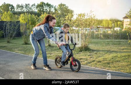 Mama hilft einem kleinen Jungen, ein Fahrrad mit zwei Rädern im Park zu fahren. Ein angenehmer Sommersporturlaub für Kinder. Stockfoto