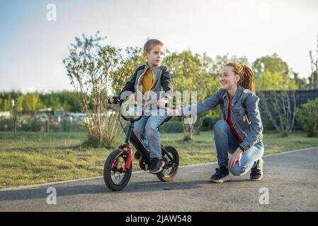 Mama hilft einem kleinen Jungen, ein Fahrrad mit zwei Rädern im Park zu fahren. Ein angenehmer Sommersporturlaub für Kinder. Stockfoto