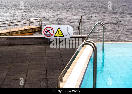 Ein Warnschild am Beckenrand des Laugarvatn Fontana-Bads in Island zeigt hier ein Sprungverbot-Symbol zusammen mit der Wassertemperatur von 32 Grad Celsius, wobei der Laugarvatn-See im Hintergrund sichtbar ist. Stockfoto