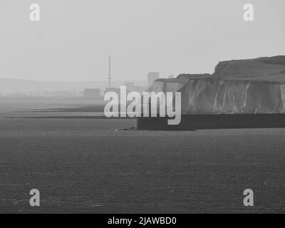 Ein dramatischer, monochromer Blick entlang einer Kreidefelsküste, mit einem Leuchtturm an einem Wellenbrecher und einer im Hintergrund verhangten Stadt. Stockfoto