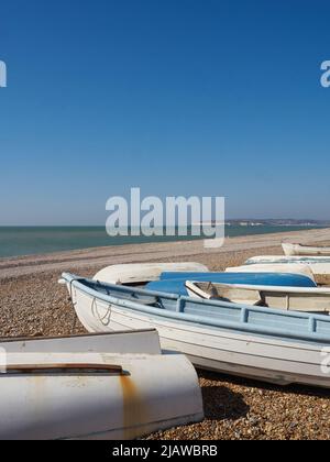 Eine Sammlung von kleinen Holzbooten, die am Kiesstrand von Seaford mit dem leeren Strand, dem Meer und der entfernten Landzunge unter einem großen, offenen Himmel auffuhren. Stockfoto