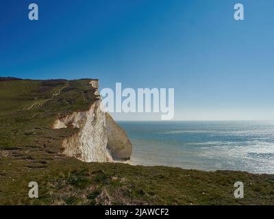 Sonnenbeschienenen weißen Kreidefelsen unter dem sommerlich blauen Himmel in der Nähe von Seaford, mit einem Pfad, der sich bis in die Ferne schlängelt und zum Erkunden einlädt. Stockfoto