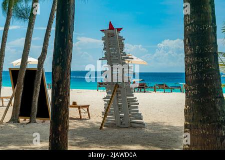 Weihnachtsbaum aus Holz und Liegestühle an einem Sandstrand vor dem Meer mit Booten, Sansibar Tansania Stockfoto