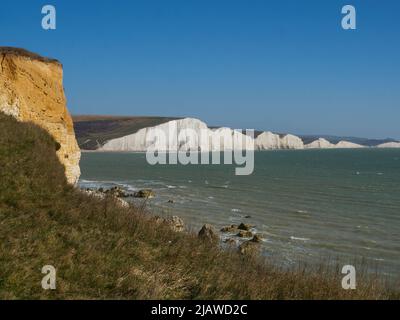 Sonnenbeschienene weiße Kreidefelsen füllen den Horizont, während die Wellen unter dem sommerlich blauen Himmel in der Nähe von Seaford an die von Felsbrocken übersäte Küste Rollen. Stockfoto