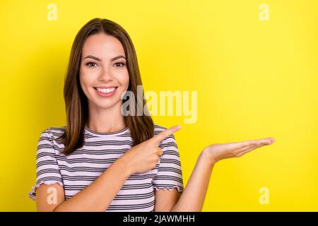 Foto von niedlichen tausendjährigen Brünette Dame Punkt halten leeren Raum tragen gestreiftes T-Shirt auf gelbem Hintergrund isoliert Stockfoto