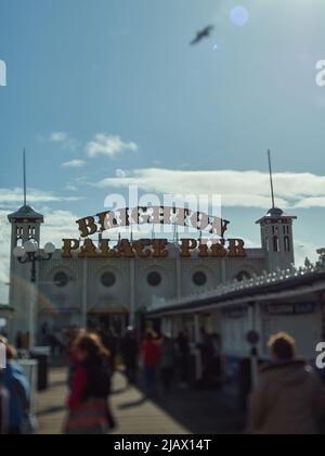Eine verschwimmerte Menge von Vergnügungsreisenden unter einem blauen, sonnenbeleuchteten Himmel geht in Richtung des berühmten Schilds, das den Weg zu den Vergnügungen am Brighton Pier zeigt. Stockfoto