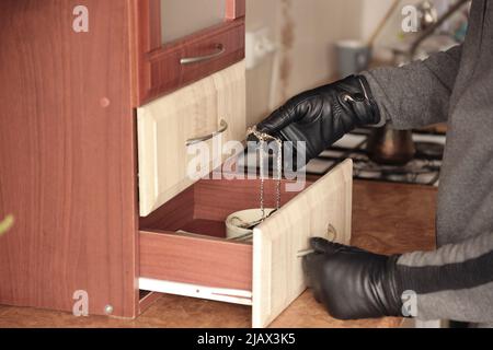 Räuber in schwarzem Outfit und Handschuhe sehen in geöffnetem Regal in der Küche. Der Dieb nimmt die goldene Halskette aus einem Küchenregal Stockfoto