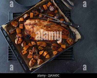 Karamellisiertes Halb-Lachsfilet mit gerösteten Kartoffeln und Kräutern auf einem Backblech serviert auf dunklem Hintergrund mit Platz zum Kopieren Stockfoto