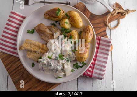 Gebratener Koalfisch mit Sauerrahm, Apfel, Zwiebel, Kräutersauce. Serviert mit gebratenen Kartoffeln auf einem Teller isoliert auf weißem Holztisch Hintergrund Stockfoto