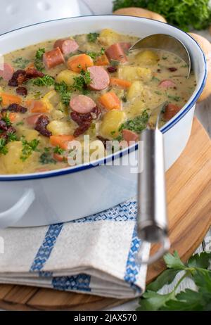 Herzhafte Suppe mit Kartoffeln, Gemüse, knusprigem Speck und gekochten Würstchen. Serviert in einem altmodischen Eintopf auf rustikalem Tischhintergrund. Stockfoto