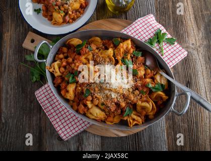 Tortellini-Gericht mit Kichererbsen, Speck in einem köstlichen Gemüse, Tomatensauce. Serviert in einer rustikalen Pfanne auf Holztisch Hintergrund mit Kopieplatz Stockfoto