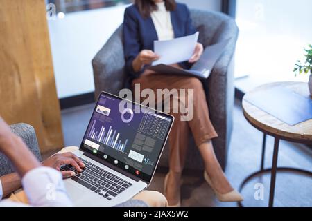 afroamerikanischer Geschäftsmann mit Laptop und kaukasischer Kollegin, die im Büro auf einem Stuhl sitzt Stockfoto