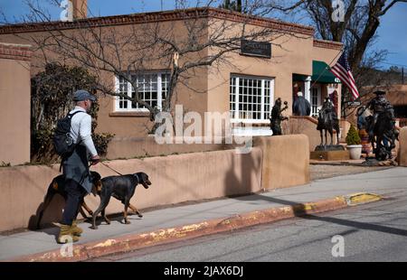 Ein Mann geht mit seinen beiden Hunden auf einem Bürgersteig vor einer Kunstgalerie in Santa Fe, New Mexico. Stockfoto