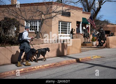 Ein Mann geht mit seinen beiden Hunden auf einem Bürgersteig vor einer Kunstgalerie in Santa Fe, New Mexico. Stockfoto