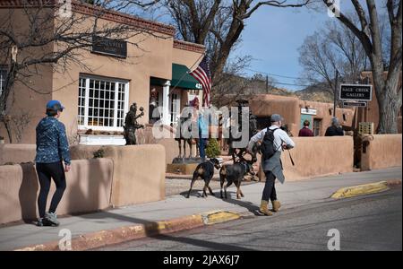 Ein Mann geht mit seinen beiden Hunden auf einem Bürgersteig vor einer Kunstgalerie in Santa Fe, New Mexico. Stockfoto