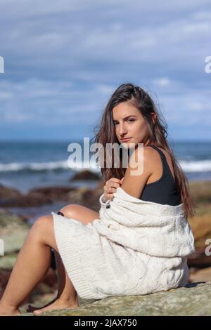 Junge Brünette Frau, die auf einem Stein sitzt und in einer beigen Strickjacke am bewölkten Strand auf die Kamera schaut Stockfoto