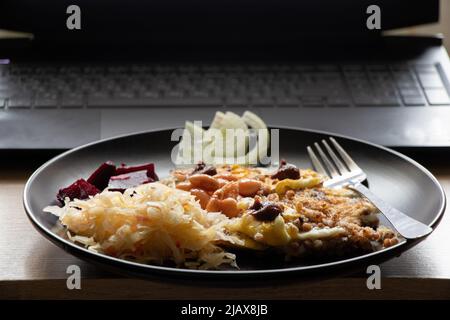 Rühreier mit Gemüse auf einem schwarzen Teller in der Nähe eines Laptops an einem Tisch auf einem Robta, Mittagessen am Arbeitsplatz, ein Teller mit Lebensmitteln am Arbeitsplatz Stockfoto