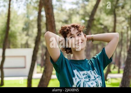 Junge Rotschopf-Frau in grünem T-Shirt auf dem Stadtpark stehend, im Freien Haare Hände hinter dem Kopf berühren und mit großem Lächeln wegschauen. Träumen von einem Stockfoto