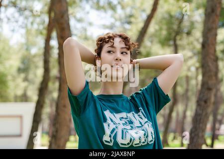 Schöne Rothaarige Frau in einem lässigen grünen T-Shirt, die auf dem Stadtpark steht und die Hände hinter dem Kopf berührt und lächelnd wegschaut. Träumen Sie abou Stockfoto