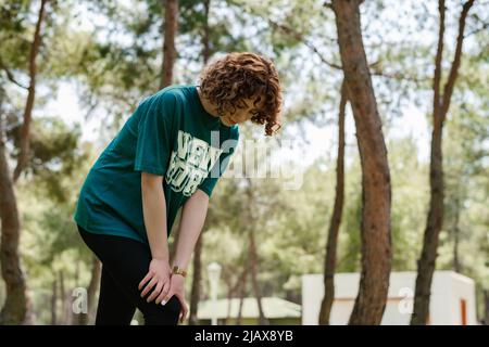 Seitenansicht einer jungen Rothaarläuferin in sportlicher Kleidung, die im Stadtpark steht und im Freien das verletzte Knie mit den Händen berührt. Verletztes Bein, gesund Stockfoto