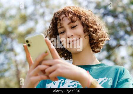 Unterseite der jungen Rothaarigen Frau, die grünes T-Shirt trägt und auf den Telefonbildschirm schaut und das Telefon benutzt. Nachrichten an Freunde senden, Videos ansehen oder scrollen Stockfoto