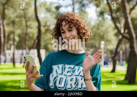 Glückliche junge Rotschopf Frau auf Stadtpark, im Freien Telefon halten Blick auf den Bildschirm winkende Hand Video Anruf Entfernung Freund online in Handy A Stockfoto
