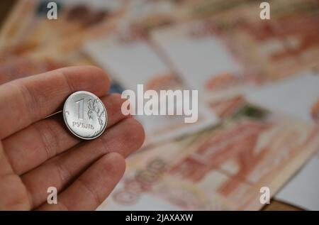 Der Mensch hält eine Eisenmünze von 1 Rubel mit der Hand auf einem verschwommenen Hintergrund von fünftausend Papierscheinen. Wirtschaft und Finanzen der Russischen Föderation. Sanktionen und Stockfoto