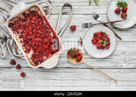 Blick von oben auf den süßen hausgemachten Erdbeer-Schuster oder Sonker, der in einer roten Keramikpfanne gebacken wird, mit einer Untertasse, einer Schürze und einem Holzlöffel. Stockfoto