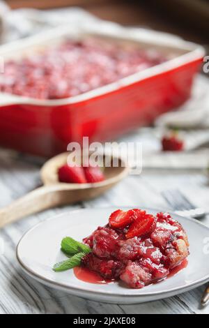 Süßer hausgemachter Erdbeer-Schuster oder Sonker, in einer roten Keramikpfanne über einem rustikalen weißen Holztisch gebacken. Extrem selektiver Fokus mit unscharfem Hintergrund Stockfoto