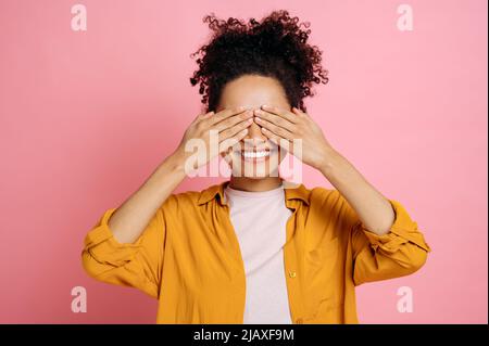 Fröhliches afroamerikanisches lockiges Mädchen in lässiger Kleidung, schließt mit den Händen die Augen, wartet auf eine Überraschung, steht auf einem isolierten rosa Hintergrund und lächelt fröhlich Stockfoto