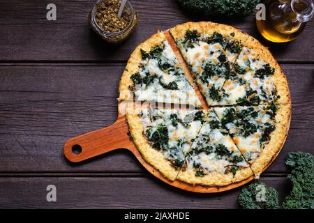 Gesunde, kohlenhydratarme Blumenkohlkruste Pizza mit Grünkohl und Pesto-Sauce. Obere Ansicht mit geschnittenen Schichten. Tischszene auf dunklem Holzhintergrund. Stockfoto