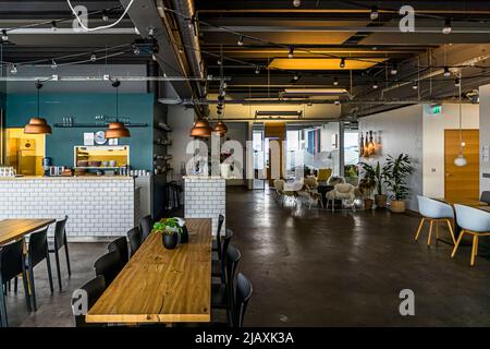 Ein offener Küchenbereich mit einer weiß gekachelten Theke und kupfernen Pendelleuchten bildet den Gemeinschaftsraum für die im Ocean Cluster House (Sjávarklasinn) ansässigen Startups der Ozeanindustrie im Hafenviertel Grandi von Reykjavík, Island. Im Hintergrund sind um einen Baumstammtisch Schafsfell-Sessel angeordnet. Stockfoto