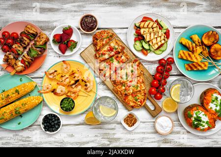 Sommer BBQ Grilltisch Szene über einem weißen Holz Hintergrund. Hähnchen- und Garnelenspieße, Fladenbrot, gefüllte Süßkartoffeln, gegrilltes Obst, Mais und Salat. Stockfoto