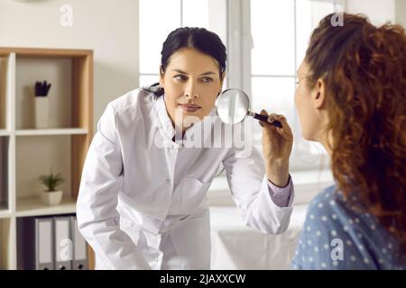 Professioneller Dermatologe mit Lupe, um die Haut auf dem Gesicht der Frau zu untersuchen Stockfoto