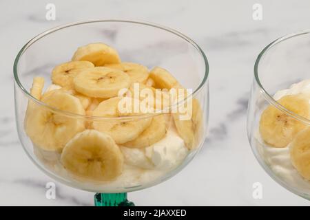 Schlagkäse-Dessert mit in Scheiben geschnittenen Bananen, serviert in Glas-Dessertbechern aus nächster Nähe auf Marmorboden Stockfoto