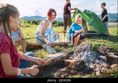 Jungen und Mädchen lachen fröhlich und rösten auf Stöcken über der Lagerfeuerflamme Marschmalchen, während zwei Brüder das grüne Zelt aufschlagen. Outdoor-Acti Stockfoto