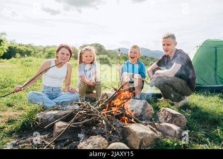 Jungen und Mädchen Kinder lachen fröhlich und rösten auf Stöcken über der Lagerfeuerflamme in der Nähe des grünen Zeltes Salmler und Würstchen. Aktive Zeit im Freien Stockfoto