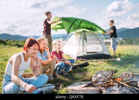 Drei Schwestern lachen fröhlich und rösten auf Stöcken über der Lagerfeuerflamme Sümpfe und Bonbons, während zwei Brüder das grüne Zelt aufschlagen. Glücklich Stockfoto