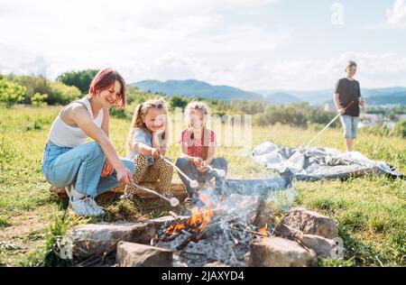 Drei Schwestern lächeln fröhlich, während sie auf den Stöcken über der Lagerfeuerflamme ein Sümmchen rösten. Fröhliche Familie oder Picknick im Freien Stockfoto