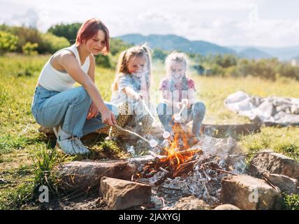 Drei Schwestern lächeln fröhlich, während sie auf den Stöcken über der Lagerfeuerflamme ein Sümmchen rösten. Fröhliche Familie oder Picknick im Freien Stockfoto