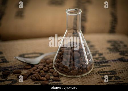 Stillleben bestehend aus einem Reagenzglas, gefüllt mit gerösteten Kaffeebohnen auf einem Sackleinen und einem Löffel daneben Stockfoto