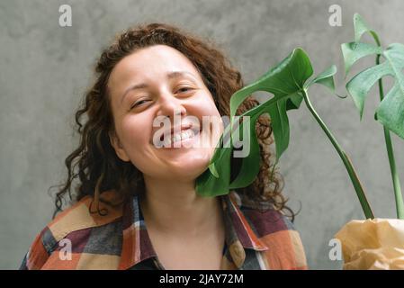 Glückliche, aufrichtig lächelnde Frau aus der Nähe sieht gerade aus und hält eine grüne Pflanze. Mädchen mit lockigen Haaren in einem karierten Hemd auf einem Hintergrund aus Betonwal Stockfoto