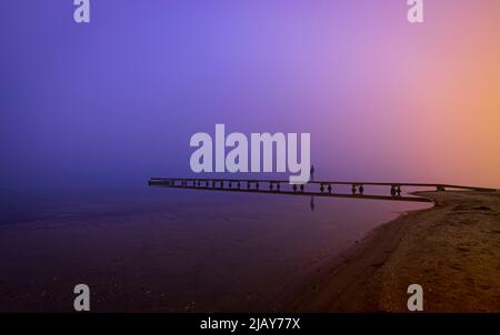 In der Abenddämmerung eine weit entfernte Silhouette einer Person auf einem hölzernen Steg mit einer geheimnisvollen Atmosphäre aus Nebel und blauen und violetten Farben. Mar Menor, Cartagena, Region Stockfoto