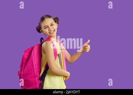 Kleines Mädchen mit Rucksack und Notizbüchern zeigt Daumen nach oben als Zeichen der Zustimmung auf lila Hintergrund. Stockfoto